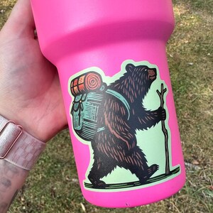 Pu&ograve; includere: Una tazza da viaggio rosa con un adesivo di un orso marrone che indossa uno zaino verde e arancione e porta un bastone. L'orso cammina su un sentiero verde.