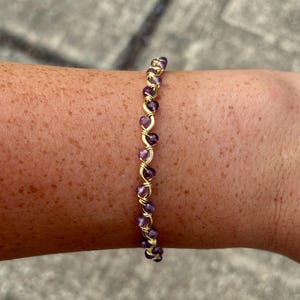 May include: A gold-colored wire bracelet with small, round, purple beads. The beads are strung together with gold wire in a spiral pattern. The bracelet is on a person's arm.