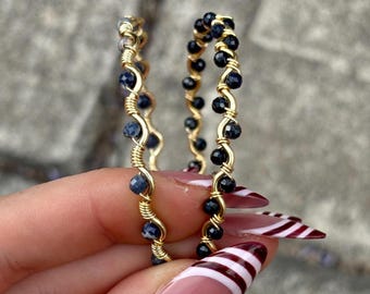 May include: Two gold-colored hoop earrings. Each earring is adorned with small, dark blue beads and wrapped with gold wire. The earrings are held up against a gray background.