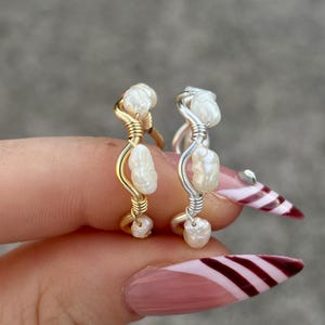 May include: Two handmade rings, one gold-toned and one silver-toned, each adorned with small, irregular white pearls. The gold ring features a twisted wire design, while the silver ring has a similar style. The rings are displayed against a blurred background.