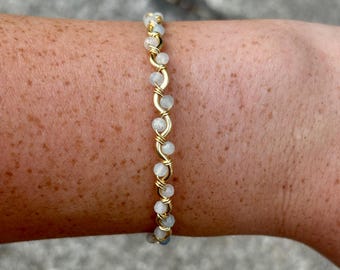 May include: A gold-colored wire-wrapped bracelet featuring small, round, light-colored beads. The beads are strung together with a delicate, interwoven design. The bracelet is shown on a person's arm.