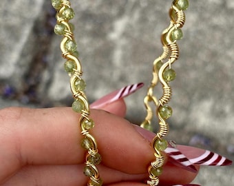May include: A pair of gold-colored hoop earrings. Each earring is adorned with small, round, light green gemstones and wrapped with gold wire. The earrings are held up against a blurred background.