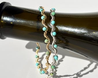 May include: Two wire-wrapped bracelets with turquoise and green beads. One bracelet is gold-toned, the other is silver-toned. The bracelets are displayed on a dark green bottle.