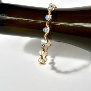 May include: A gold-colored wire-wrapped bracelet with small, round, opalescent beads. The bracelet is curved and rests on a dark brown surface, with a shadow cast below. The beads are evenly spaced along the wire.