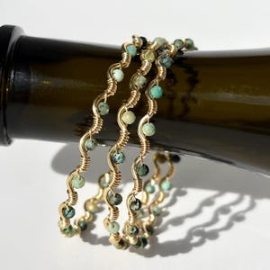 Op de afbeelding: Vier goudkleurige draadarmbanden met een golvend ontwerp, elk geregen met kleine, ronde, groene en turquoise kralen. De armbanden worden gepresenteerd op een donkergroene fles, met een witte achtergrond.