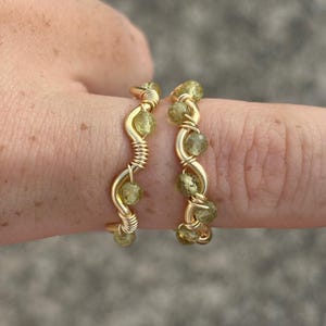 Könnte beinhalten: Zwei goldfarbene Ringe mit Drahtwickeldesign. Jeder Ring hat kleine, runde, hellgrüne Perlen. Die Ringe werden auf einem Finger vor einem verschwommenen, neutralen Hintergrund präsentiert. Die Ringe haben ein zartes, handgefertigtes Aussehen.