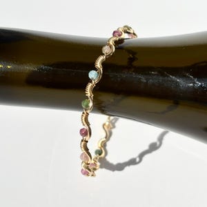May include: A gold-coloured wire-wrapped bracelet featuring a series of colourful gemstones. The gemstones are in shades of pink, green, and blue. The bracelet is curved and rests on a dark surface.