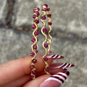 May include: Two gold-coloured hoop earrings, each adorned with small, round, deep red gemstones. The gemstones are evenly spaced and wrapped with gold wire. The earrings are held up against a blurred background.