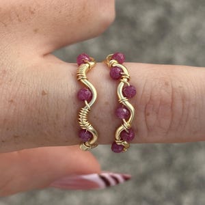 May include: Two gold-colored wire rings, each adorned with small, round, deep pink gemstones. The rings have a wavy design, with the gemstones spaced along the curves. The rings are displayed on a finger.