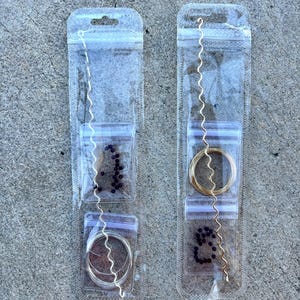 May include: Two clear plastic packages containing jewelry-making supplies. One package has silver-colored wire, beads, and a wavy silver wire. The other has gold-colored wire, beads, and a wavy gold wire. The packages are on a gray surface.