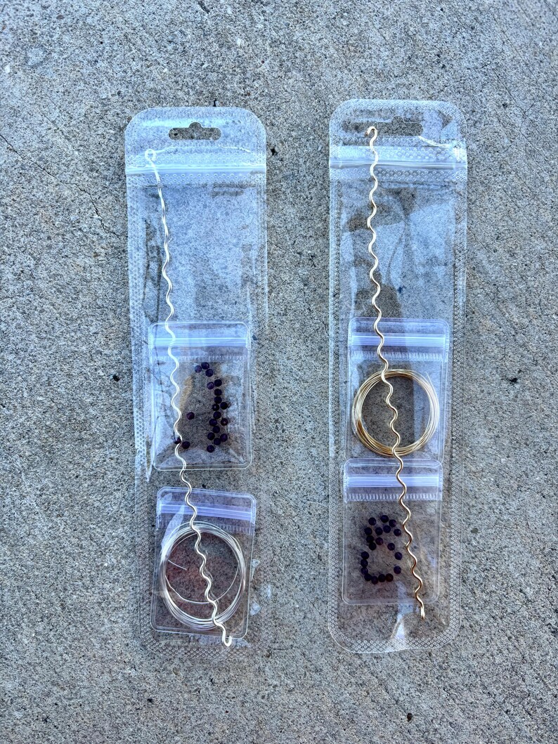 May include: Two clear plastic packages containing jewelry-making supplies. One package has silver-colored wire, beads, and a wavy silver wire. The other has gold-colored wire, beads, and a wavy gold wire. The packages are on a gray surface.