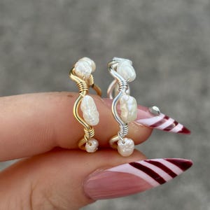 May include: Two handmade rings with freshwater pearls. One ring is gold-toned, the other is silver-toned. The pearls are clustered and wrapped with wire. The rings are held up by a hand with a pink manicure.