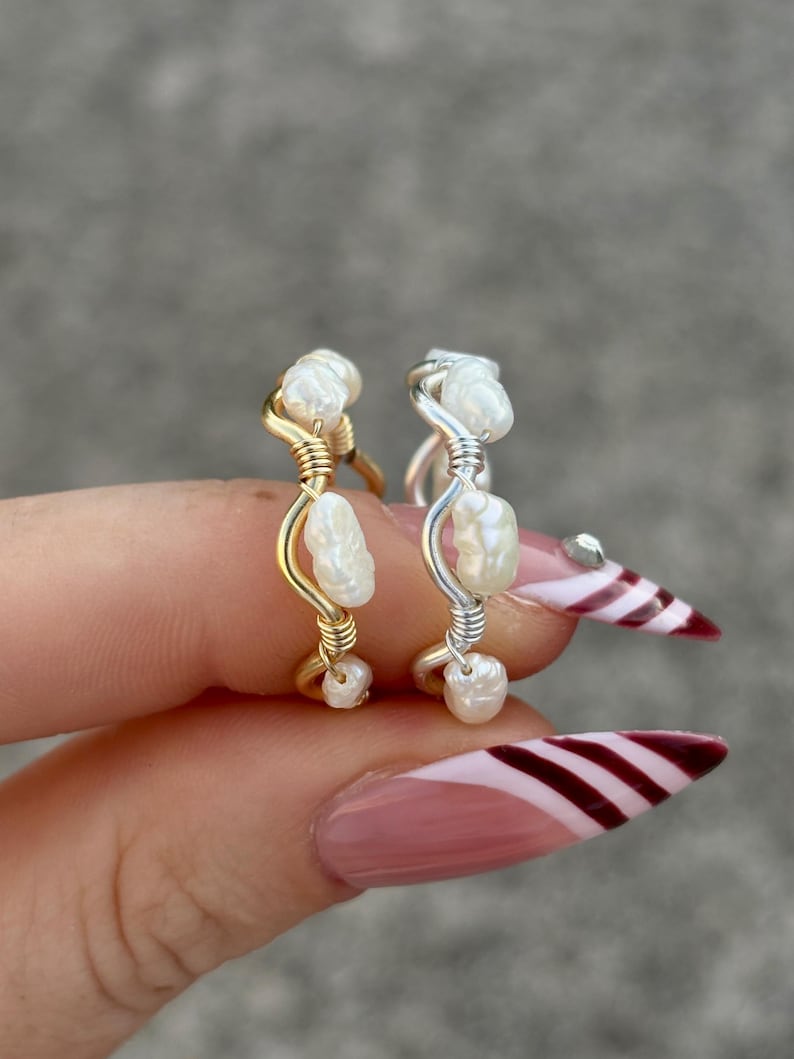 May include: Two handmade rings with freshwater pearls. One ring is gold-toned, the other is silver-toned. The pearls are clustered and wrapped with wire. The rings are held up by a hand with a pink manicure.