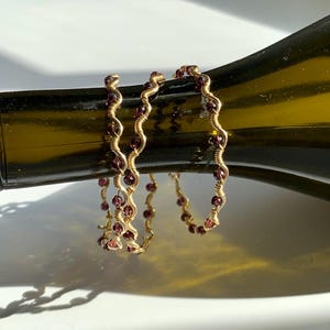 May include: Gold-coloured wire bracelets with a wavy design, decorated with small, round, red gemstones. The bracelets are displayed on a dark green glass bottle, creating a contrast of colours and textures.