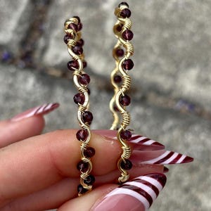 May include: A pair of gold-colored hoop earrings. Each earring is adorned with dark red beads and wrapped with gold wire. The earrings are displayed against a blurred background.
