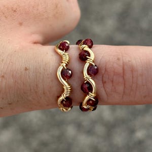 Könnte beinhalten: Zwei goldfarbene Drahtringe, jeweils verziert mit kleinen, runden, tiefroten Edelsteinen. Die Ringe werden auf einem Finger präsentiert und zeigen die komplizierte Drahtwicklung und die leuchtende Farbe der Edelsteine. Der Hintergrund ist verschwommen und neutral.