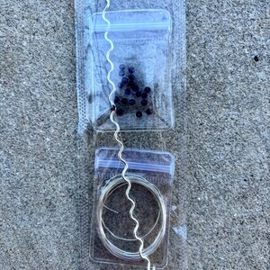 May include: A jewelry-making kit featuring a silver-colored, wavy wire, a small bag of dark purple beads, and a coil of silver wire, all packaged in clear plastic bags. The kit is designed for crafting necklaces, bracelets, or other jewelry.