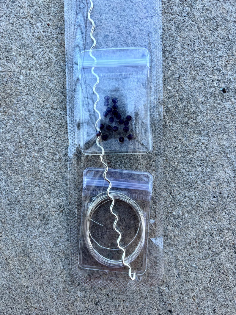 May include: A jewelry-making kit featuring a silver-colored, wavy wire, a small bag of dark purple beads, and a coil of silver wire, all packaged in clear plastic bags. The kit is designed for crafting necklaces, bracelets, or other jewelry.