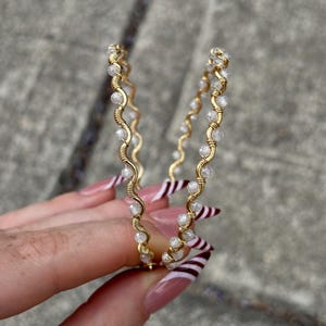 May include: A pair of gold hoop earrings. Each earring has a wavy design with small, clear beads and wrapped wire detailing. The earrings are held against a grey background.