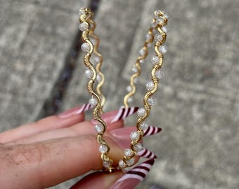 May include: A pair of gold-colored hoop earrings. Each earring features a wavy design with small, clear beads and wrapped wire detailing. The earrings are held up against a gray background.