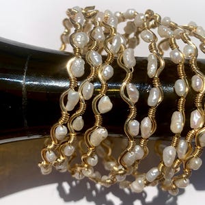 May include: Several gold-toned bracelets with freshwater pearls. The bracelets are made with gold wire wrapped around the pearls in a wavy pattern. The pearls are white and irregularly shaped. The bracelets are stacked together.