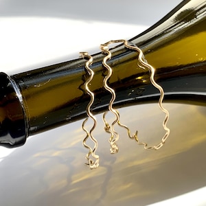 以下が含まれることがあります： 金色の波型のフープピアス3つ。ピアスは濃い緑色のガラス瓶に飾られています。ピアスは繊細でミニマリストなデザインで、連続した流れるような形をしています。