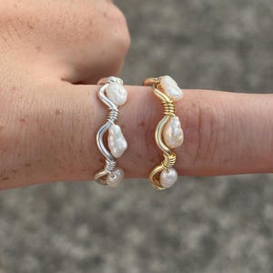 May include: Two rings with freshwater pearls, one silver-toned, the other gold-toned. Each ring has three white pearls. The rings are wire-wrapped. The rings are displayed on a finger against a blurred background.