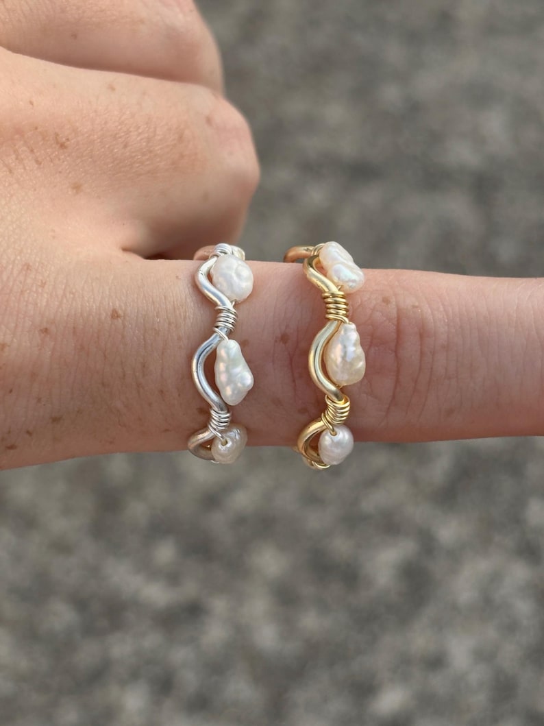 May include: Two rings with freshwater pearls, one silver-toned, the other gold-toned. Each ring has three white pearls. The rings are wire-wrapped. The rings are displayed on a finger against a blurred background.