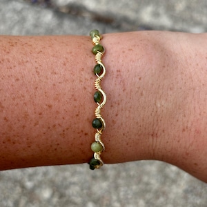 May include: A gold-colored wire-wrapped bracelet featuring green beads. The beads vary in shade from light to dark green. The bracelet is designed to be worn on the wrist and is displayed against a skin background.
