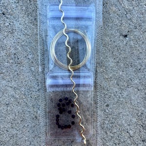 May include: A clear plastic package contains jewelry-making supplies. Inside are a coil of gold wire, a wavy gold wire, and a small pile of dark purple beads. The package is sealed with a zip-top closure.