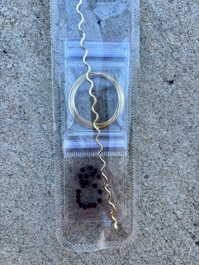 May include: A clear plastic package contains jewelry-making supplies. Inside are a coil of gold wire, a wavy gold wire, and a small pile of dark purple beads. The package is sealed with a zip-top closure.