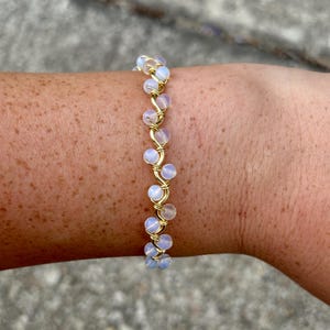 Könnte beinhalten: Ein filigranes, goldfarbenes Drahtarmband mit einer Reihe kleiner, schillernder, runder Perlen. Die Perlen sind von weicher, milchig-weißer Farbe mit einem dezenten Blaustich. Das Armband ist für das Handgelenk konzipiert.