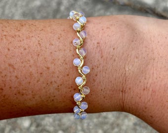 May include: A delicate gold-toned wire bracelet featuring a series of small, iridescent, round beads. The beads are a soft, milky white color with a subtle blue hue. The bracelet is designed to be worn on the wrist.
