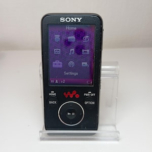 Puede incluir: Reproductor de música digital Sony Walkman negro con una pantalla púrpura que muestra iconos y texto. El dispositivo tiene un botón de control circular y el logotipo de Sony. Se sostiene en un soporte de plástico transparente.