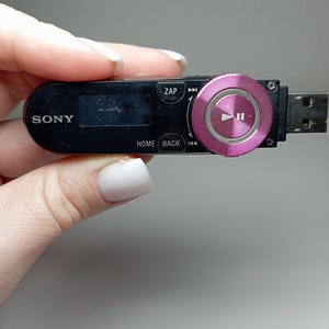 May include: Black Sony MP3 player with a pink control wheel and USB connector. The player has a small screen and buttons labeled "ZAP", "HOME", and "BACK". The control wheel has a play/pause symbol.