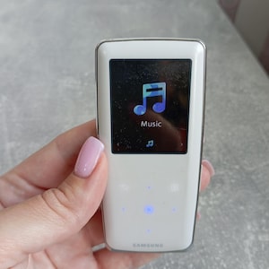 May include: A white Samsung digital music player with a rectangular screen displaying a blue musical note icon and the word Music. The device features a smooth, glossy finish with a touch-sensitive control panel below the screen and a silver trim.