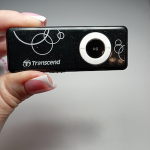 May include: Black Transcend MP3 player with white circular design elements. The player has a white control dial with play/pause and forward/rewind buttons. The Transcend logo is visible on the device.