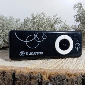 May include: A black rectangular Transcend digital music player featuring a white circular navigation pad with play, pause, and track control icons. The device surface is decorated with thin white line art of overlapping circles and the Transcend logo.