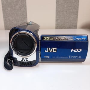 Puede incluir: Una videocámara de disco duro JVC Everio de color azul oscuro con lente Konica Minolta. El panel lateral muestra texto que incluye 30GB, 7 to 37 HRS, 4 Rec Modes, Hybrid, JVC, HDD y Micro SD. El dispositivo cuenta con una correa negra y un anillo de lente plateado.