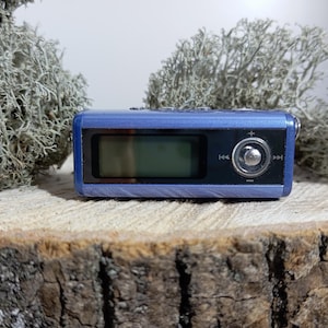 May include: A rectangular blue digital music player featuring a black front panel with a small screen, a circular navigation button in the center, and playback control icons including skip forward, skip backward, volume plus, and volume minus.