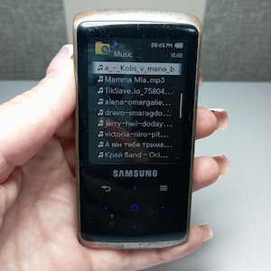 May include: A black Samsung MP3 player with a silver frame is held in a hand. The screen displays a music playlist with song titles. The time is 8:26 PM, and the date is 10/23. The player has a few control buttons.