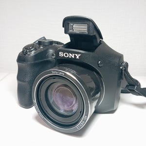 May include: Black Sony digital camera with a pop-up flash and a lens that reads "Sony Lens". The camera has a 26x optical zoom and the lens reads 3.1-5.9/4-104. The camera has a black strap attached.