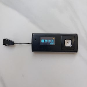 May include: A black MP3 player with a small screen displaying icons and text. The device has a large play/pause button and a connected USB cable. The screen shows the time as 13:58.