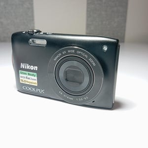 May include: A black Nikon Coolpix digital camera. It features a slim body, wide 6x zoom, and 16.0 megapixels. The lens is marked with "NIKKOR 6X WIDE OPTICAL ZOOM" and "4.6-27.6mm 1:3.5-6.5". The camera is on a white surface.
