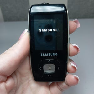 May include: Black Samsung digital media player with a glossy finish. The screen displays the Samsung logo. The device has a central control button and arrow keys. The device is held in a hand.