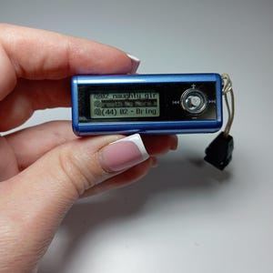Può includere: Un piccolo lettore MP3 blu con uno schermo che mostra i titoli delle canzoni. Lo schermo mostra "02 naughty girl" e "(44) 02 - Bring". Il lettore ha un pulsante di controllo argentato e un accessorio nero.