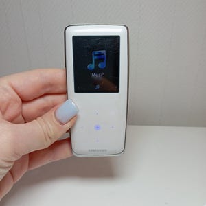 May include: White Samsung MP3 player with a black screen showing a blue music note icon and the word "Music". The device has a silver trim and a blue light. The Samsung logo is visible.
