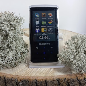 May include: A black Samsung portable media player with a silver frame and a digital screen displaying icons for music, videos, pictures, texts, FM radio, datacasts, prime pack, file browser, and settings, with the current time shown as 03:44 PM.