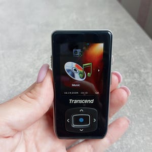 Puede incluir: Un reproductor de música digital Transcend negro con una pantalla rectangular que muestra un icono de música, la fecha 19.02.2026 y la hora 12:12. El dispositivo cuenta con un panel de navegación central con flechas y un botón azul.