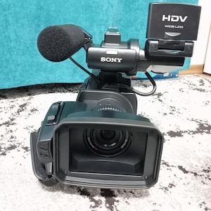 Puede incluir: Una cámara de vídeo Sony HDV negra con micrófono y pantalla LCD ancha. La cámara tiene una lente con las marcas 1.8/5.4-58. La cámara está sobre una alfombra de color claro.